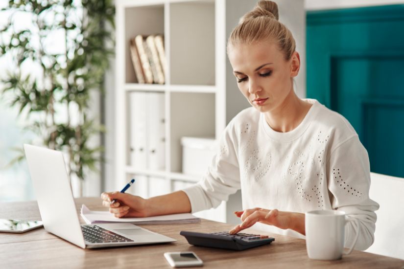 Subtract your expenses from your income. A woman sitting at a desk using a calculator and a laptop.